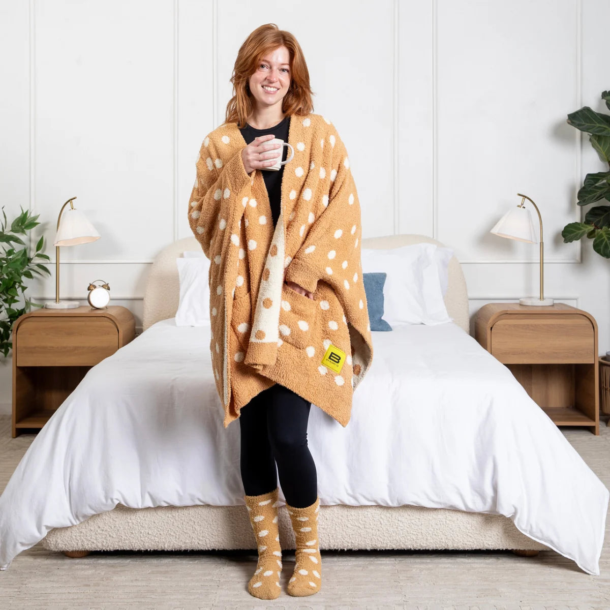 Woman wearing tan oversized blanket wrap with white polka dots, holding mug on bed