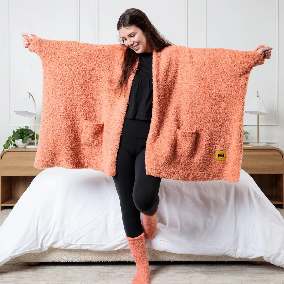 Woman wearing oversized coral melon textured plush wrap blanket with pockets on bed