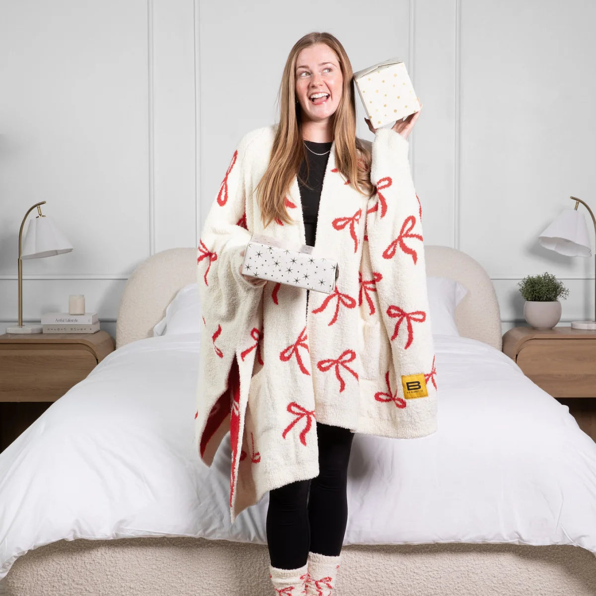 Woman wearing cream oversized blanket wrap with red bow pattern, holding coordinating throw pillow on bed