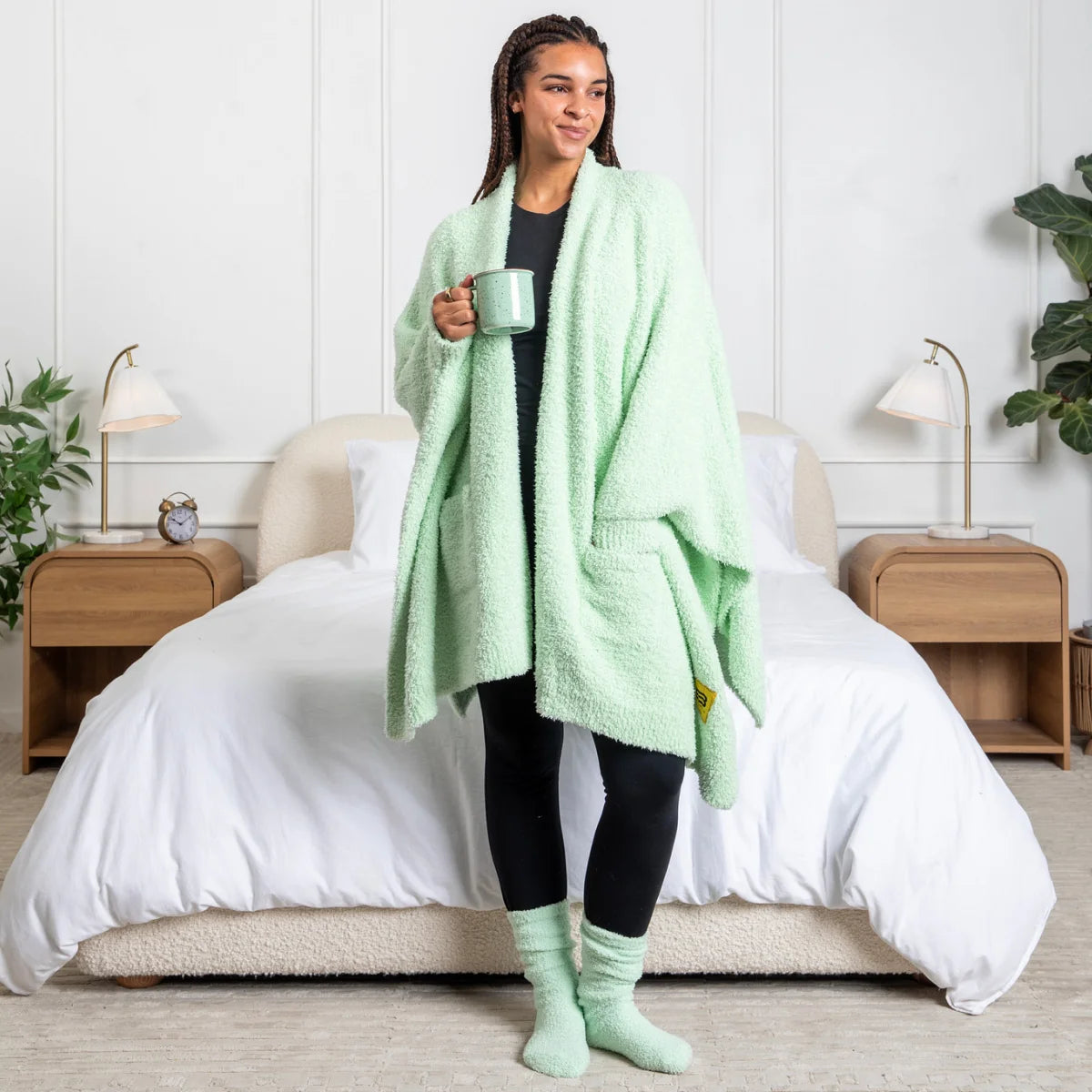 Woman wearing mint green oversized blanket wrap draped over shoulders while holding mug on bed