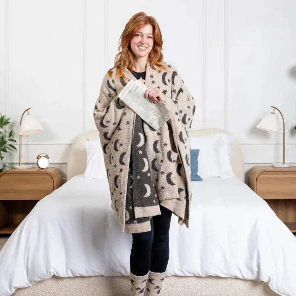 Woman wearing cream and charcoal celestial-print oversized blanket wrap over black clothing, standing on bed with white pillows