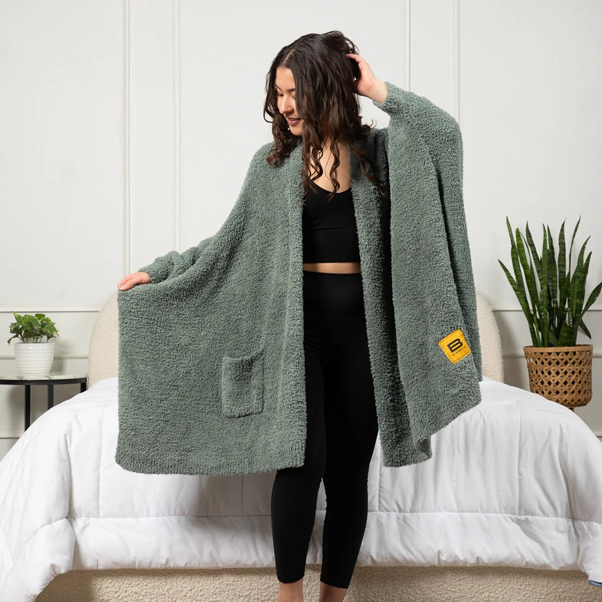 Woman wearing moss-green oversized blanket wrap with pockets draped over shoulders while sitting on bed