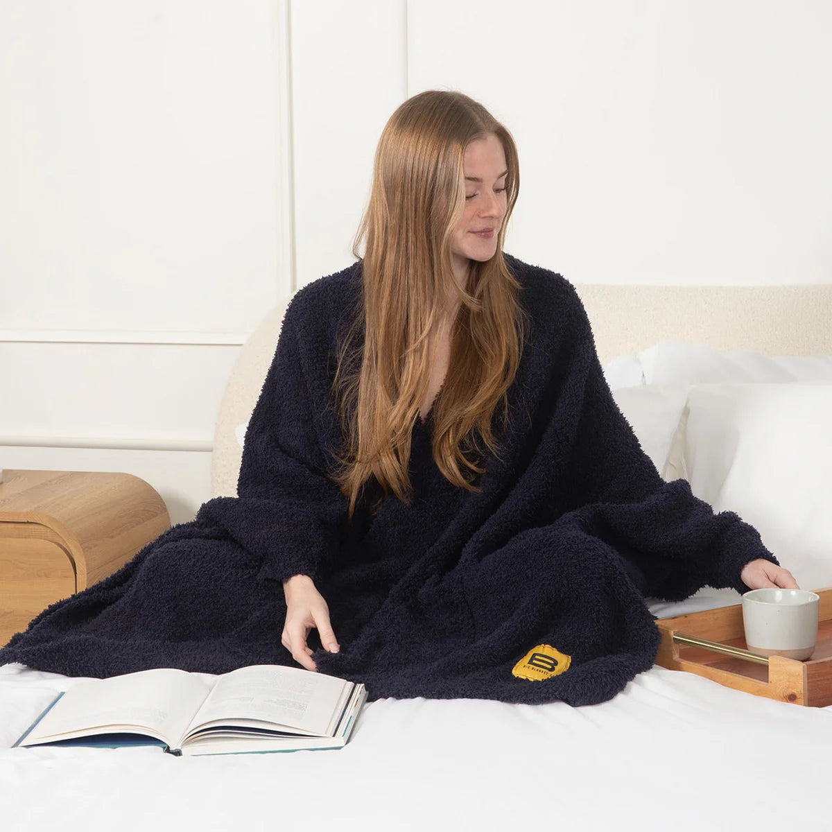 Woman wrapped in navy oversized blanket while reading on bed with coffee cup and book