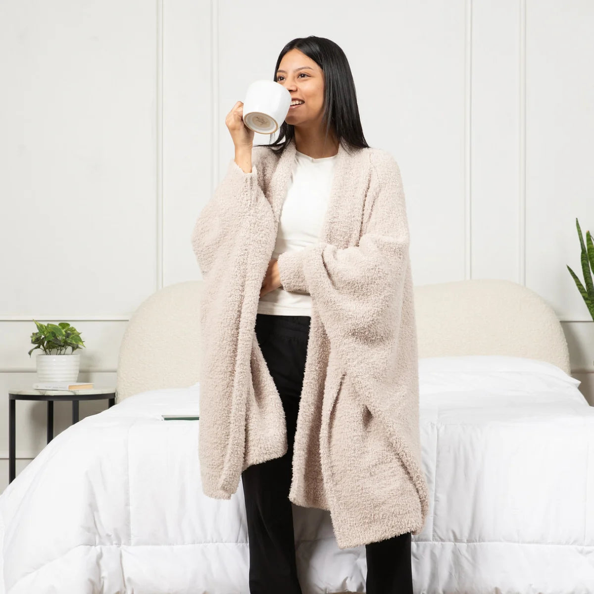 Woman wearing oatmeal Premier Plush Wrap oversized blanket cardigan over white shirt while drinking from mug on bed