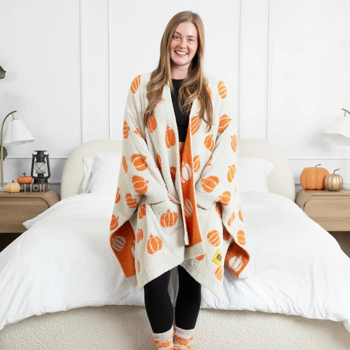 Woman wearing cream and orange pumpkin print oversized blanket wrap on bed with fall decor