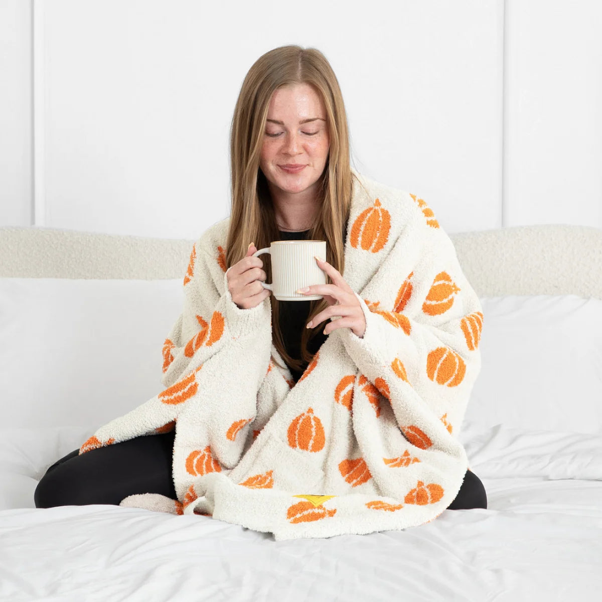 Woman wrapped in cream oversized blanket with orange pumpkin pattern, holding mug on white bed