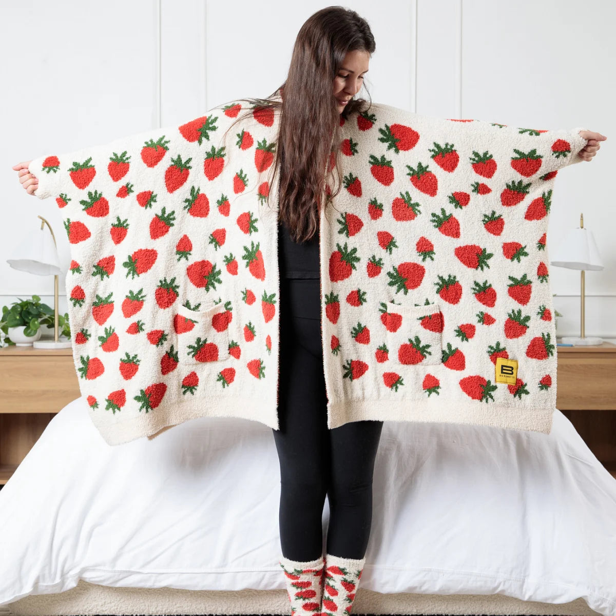 Woman holding oversized cream blanket with red strawberry and green leaf pattern draped across shoulders on bed