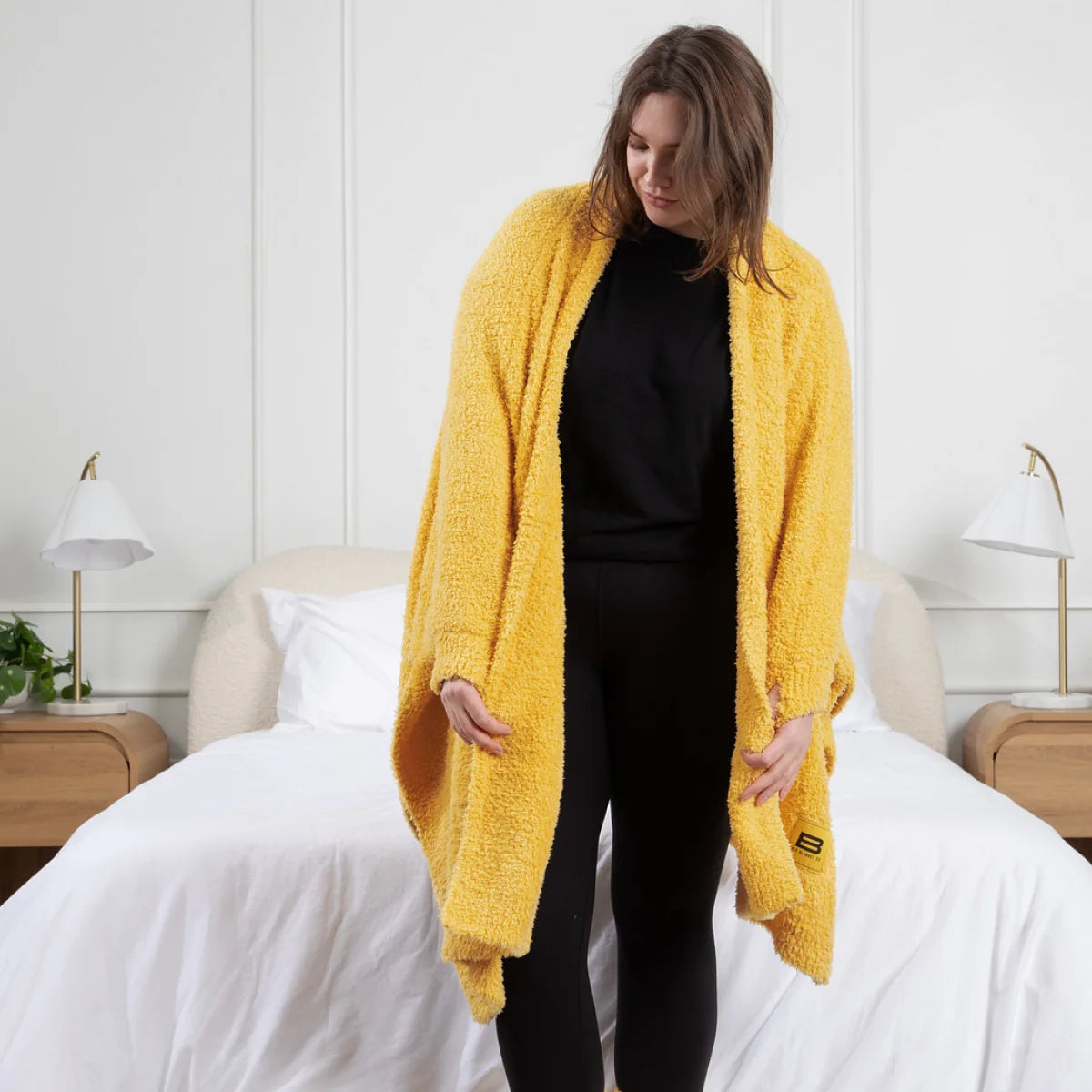 Woman wearing oversized golden yellow textured blanket wrap draped over shoulders on bed with white bedding