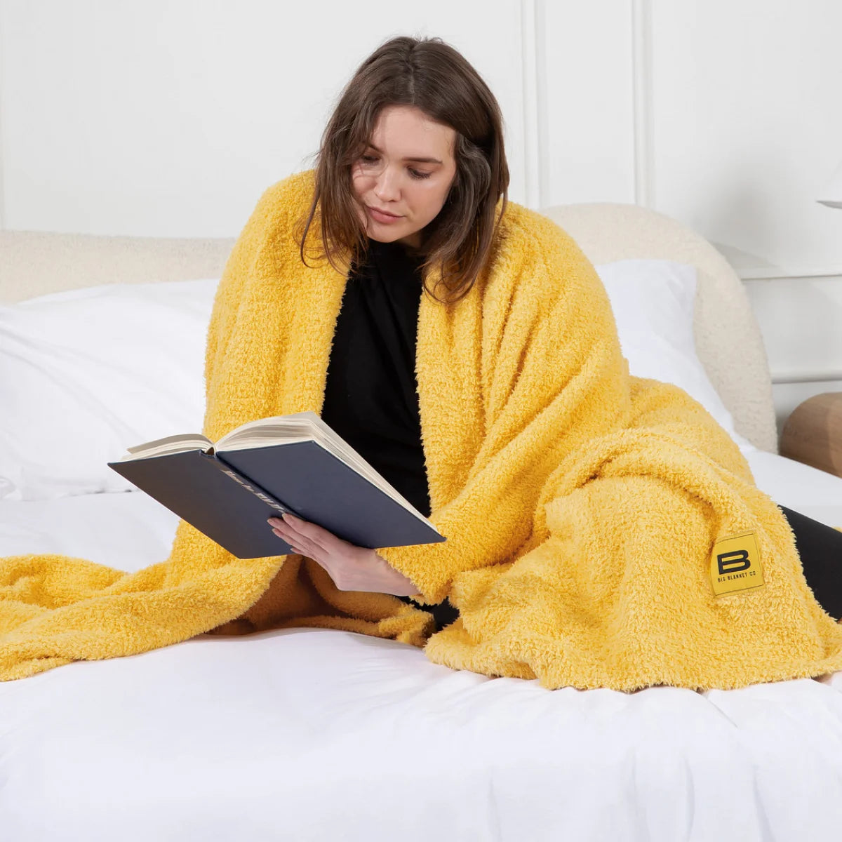 Woman wrapped in yellow oversized blanket reading on bed