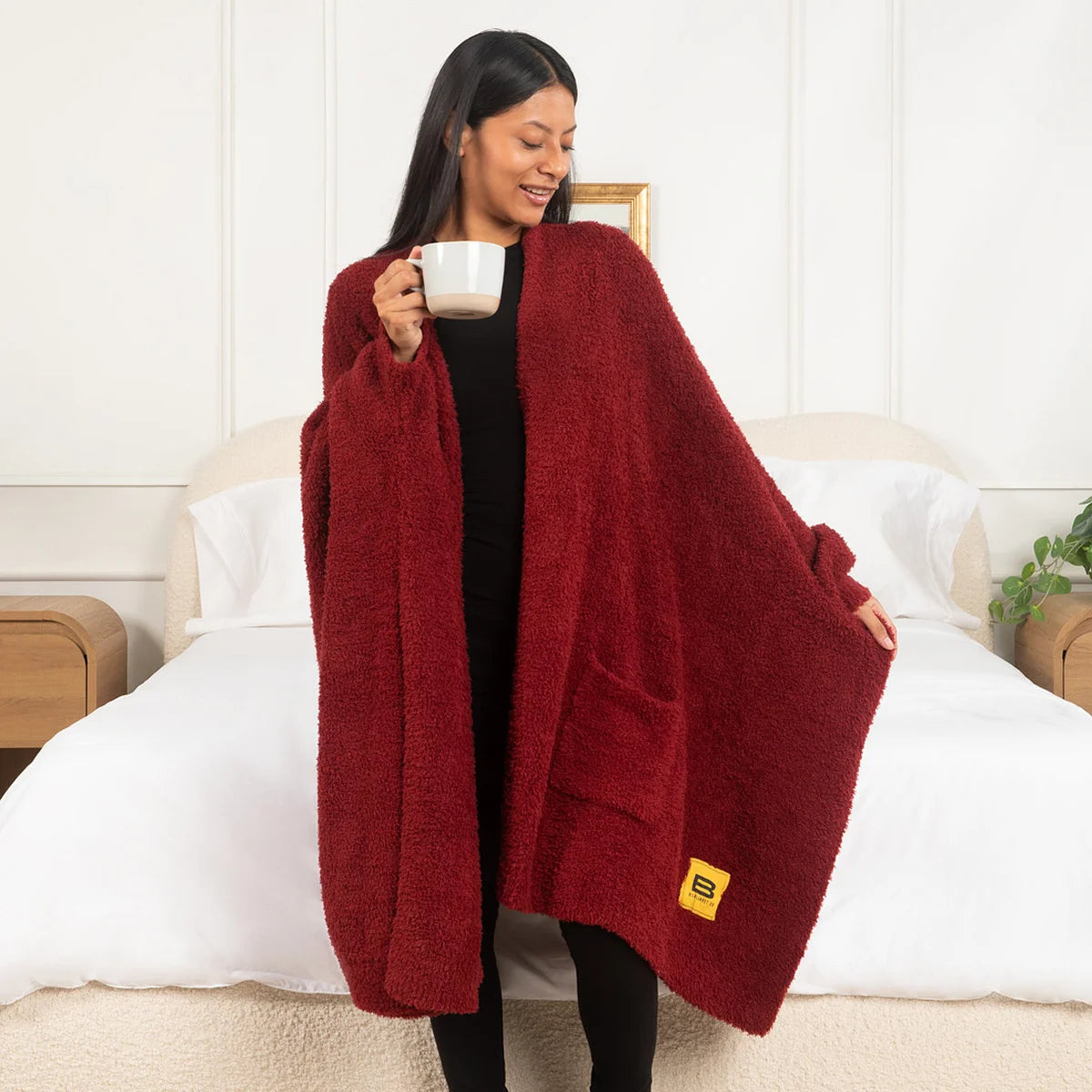 Woman wrapped in burgundy oversized blanket sitting on bed holding mug