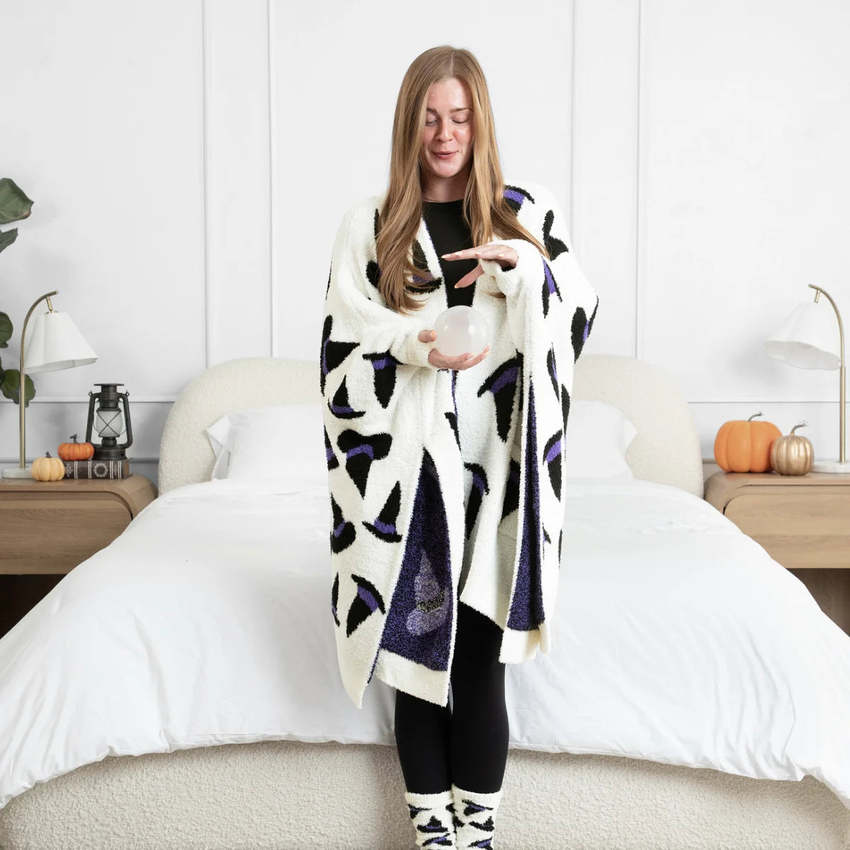 Woman wearing cream oversized blanket wrap with black and purple geometric pattern, standing on bed in modern bedroom