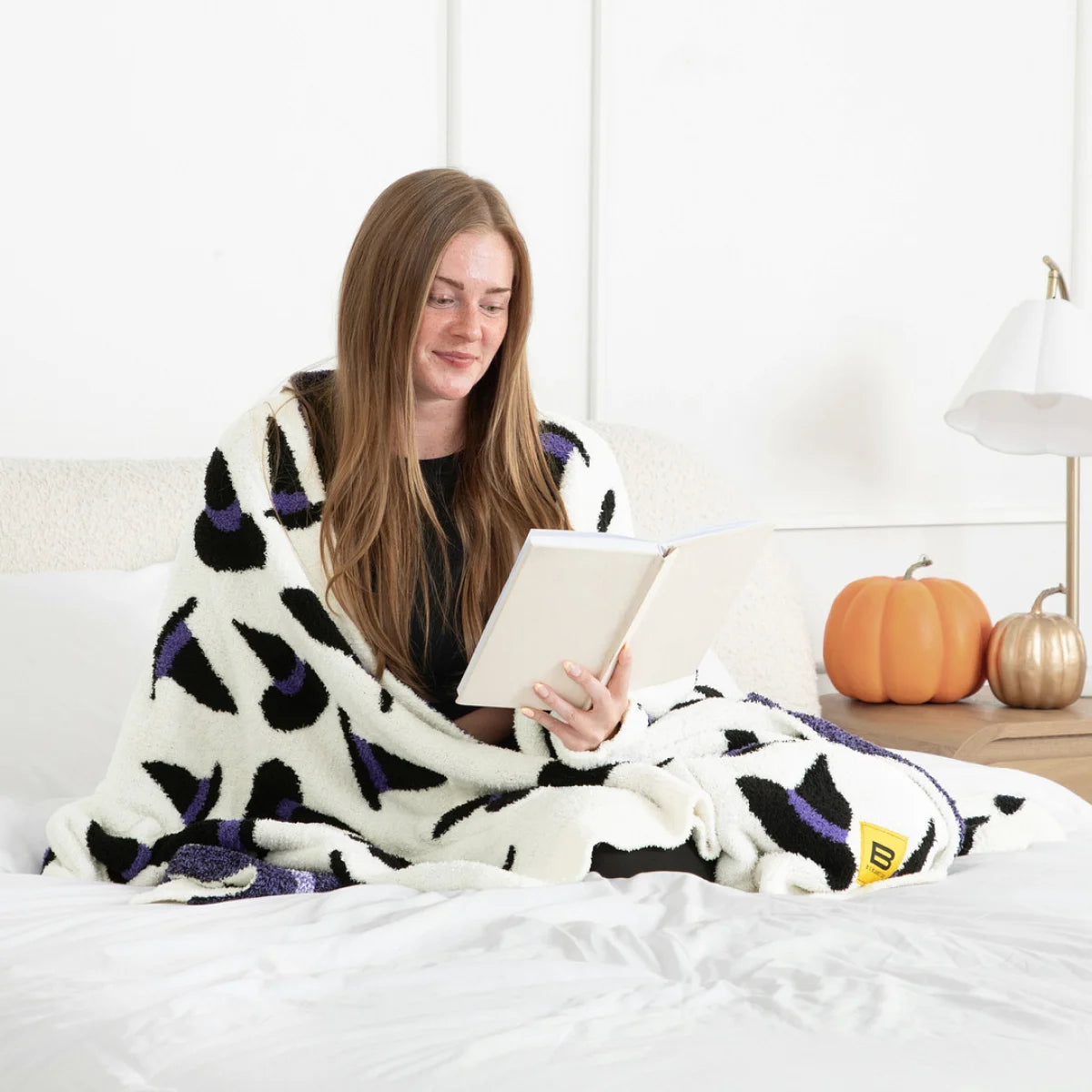 Woman wrapped in oversized cream blanket with black and purple geometric pattern, reading tablet on bed